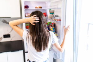 An Eating Disorder vs disordered eating could look like a woman standing at open refrigerator looking overwhelmed.